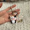 A handmade crochet cow keychain featuring a white and black cow figure with pink accents and gold hardware.