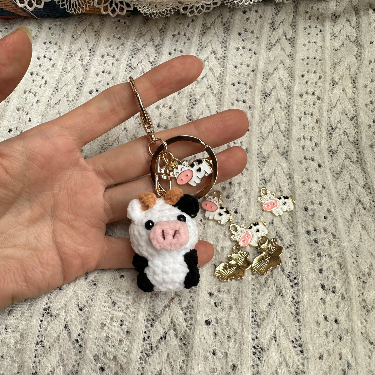 A handmade crochet cow keychain featuring a white and black cow figure with pink accents and gold hardware.