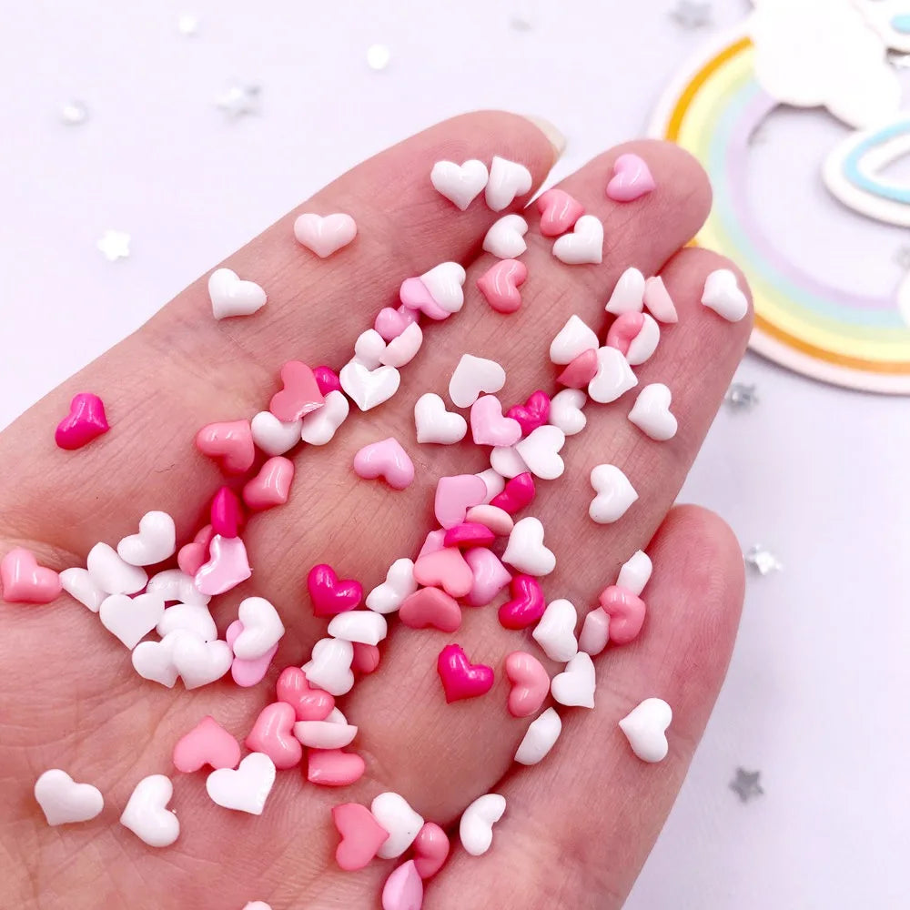A hand holds a mix of pink and white heart-shaped flatback rhinestones for nail art and crafts.