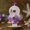 A white birthday candle shaped like the number eight decorated with purple glitter and a silver starfish flower.