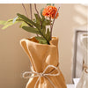 A beige ceramic money bag shaped vase holding an orange flower, displayed on a white surface.