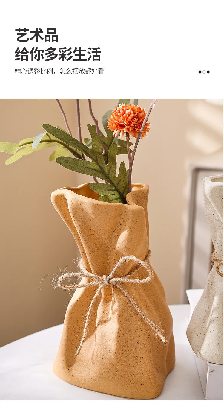 A beige ceramic money bag shaped vase holding an orange flower, displayed on a white surface.
