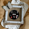 A set of black button pins featuring punk band album cover designs and a butterfly decoration displayed in a white frame.