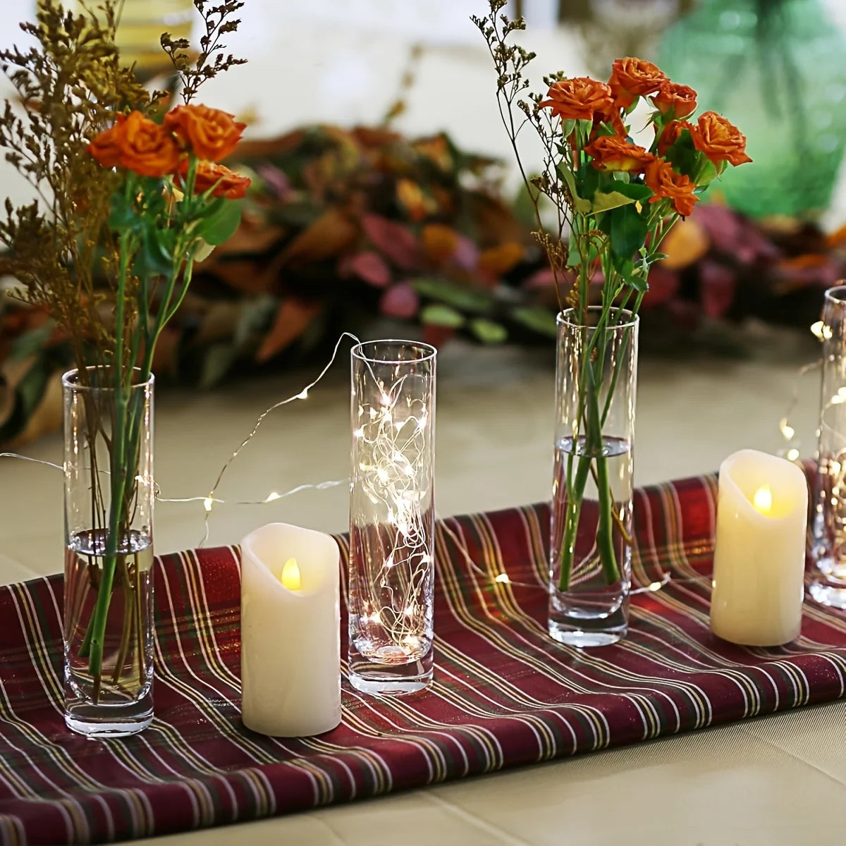 A set of clear glass cylinder vases with dried flowers and fairy lights arranged on a red fabric surface.