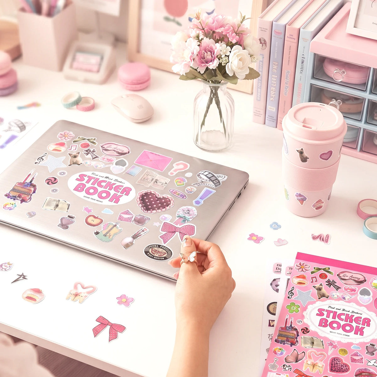 A hand holds a pink aesthetic sticker book with decorative stickers on a white table next to floral arrangements and statione