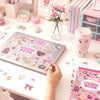 A hand holds a pink aesthetic sticker book with decorative stickers on a white table next to floral arrangements and statione