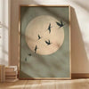 A framed canvas wall art featuring a large pale circle resembling a moon with several dark silhouettes of cranes flying acros