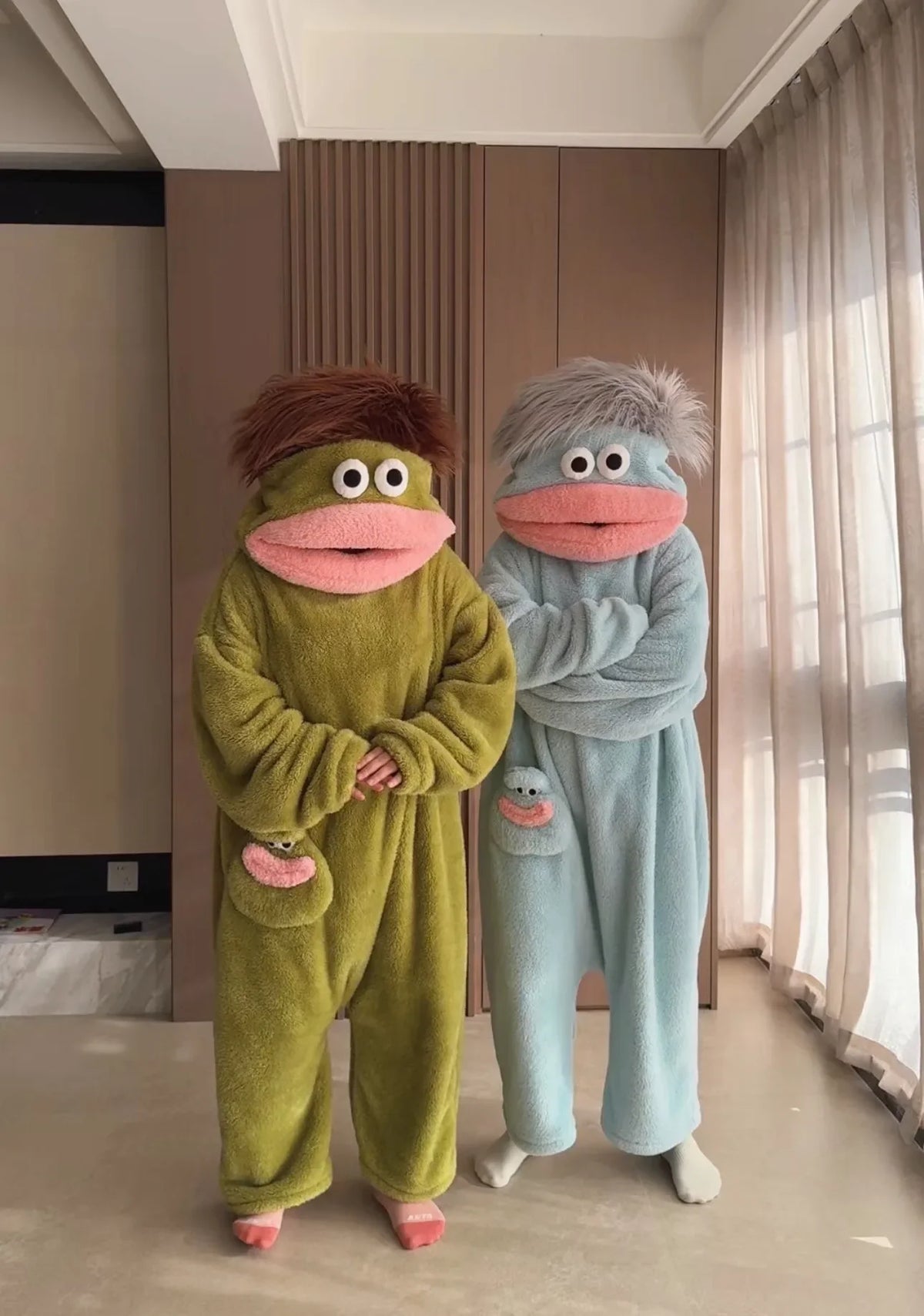 A pair of fuzzy green and blue monster onesie pajamas for couples standing in a room.