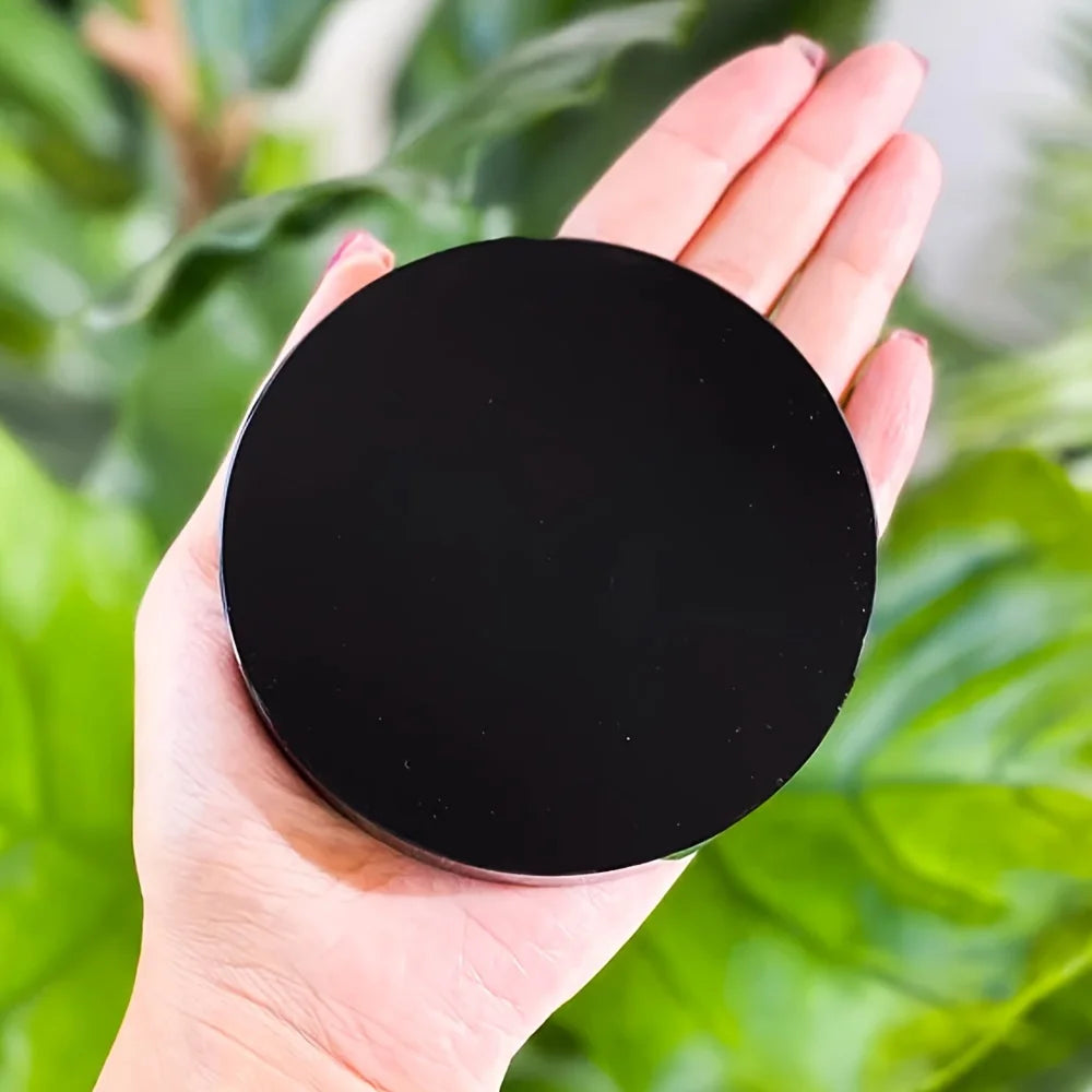 A hand holds a round, black obsidian mirror used for Feng Shui and divination against a blurred green background.