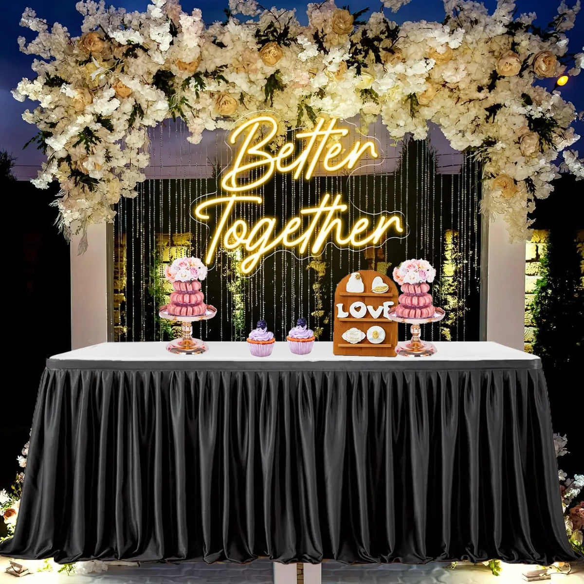 A black pleated polyester table skirt displayed at an event setup with floral decor and desserts.
