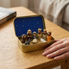 A hand-painted mini nativity set in a gold tin storage box resting on a wooden table.