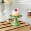 A mini green cupcake stand displaying a decorated cupcake with cream and a strawberry on a wooden surface.