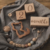 A collection of wooden baby milestone cards and a wooden silhouette necklace displayed on a gray linen cloth.