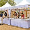 A white table draped with vertical pleats is set up under a large tent for an event.
