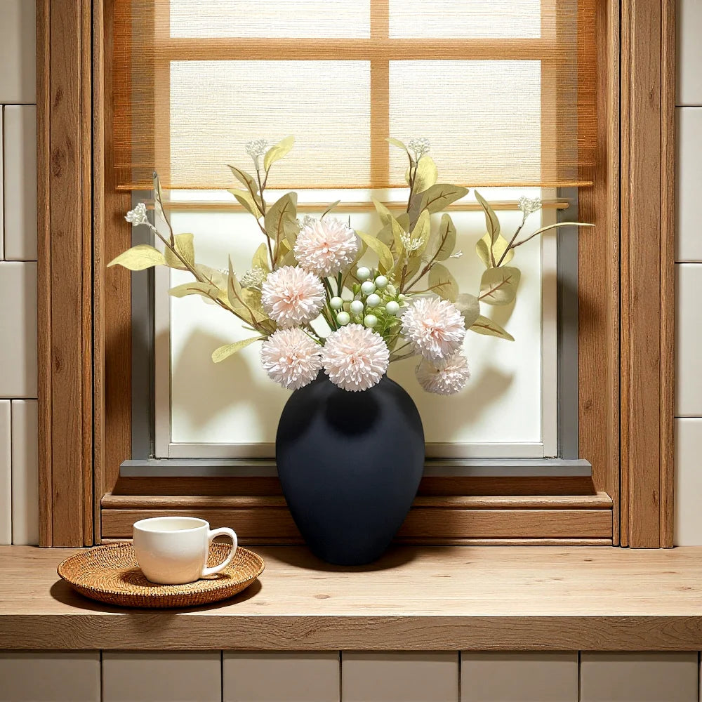 A dark ceramic vase filled with light pink and green floral arrangements sits on a wooden shelf near a window.