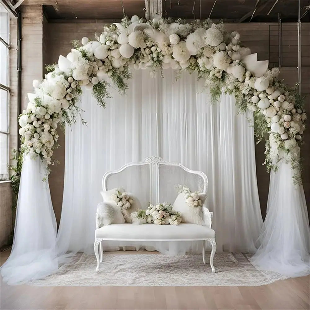 A wedding arch decorated with white flowers and greenery, framing a white bench and sheer fabric backdrop.