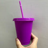 A hand holds a bright purple plastic cold cup with a pink straw against a light gray wall.
