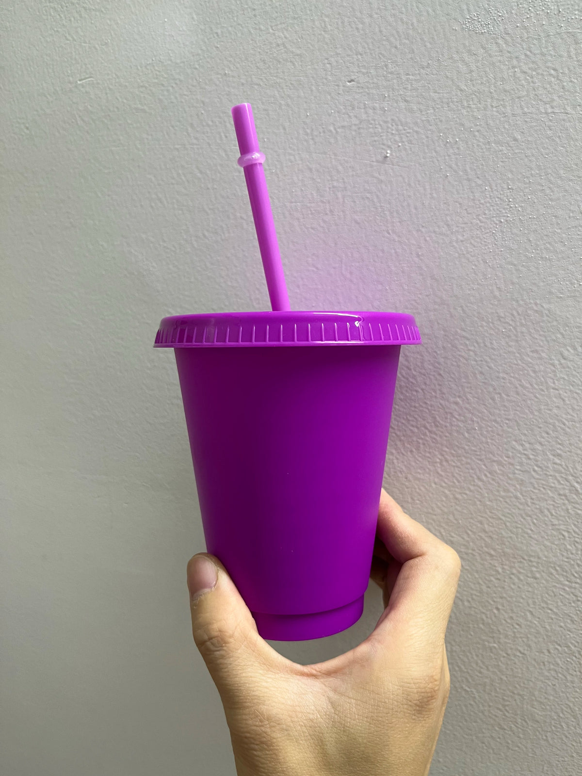 A hand holds a bright purple plastic cold cup with a pink straw against a light gray wall.
