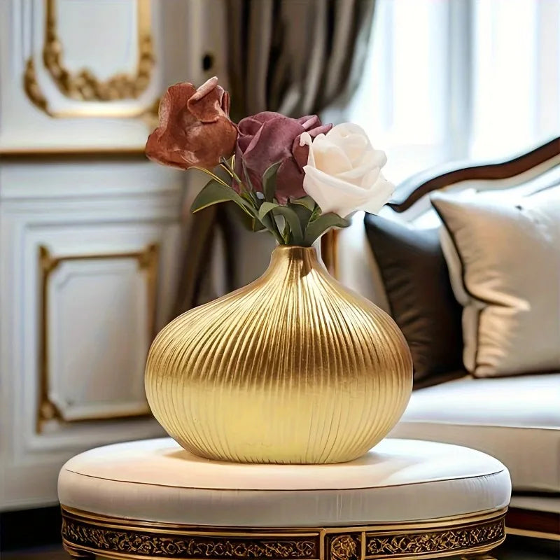 A golden striped resin vase with deep pink and white roses sits on a round white pedestal.