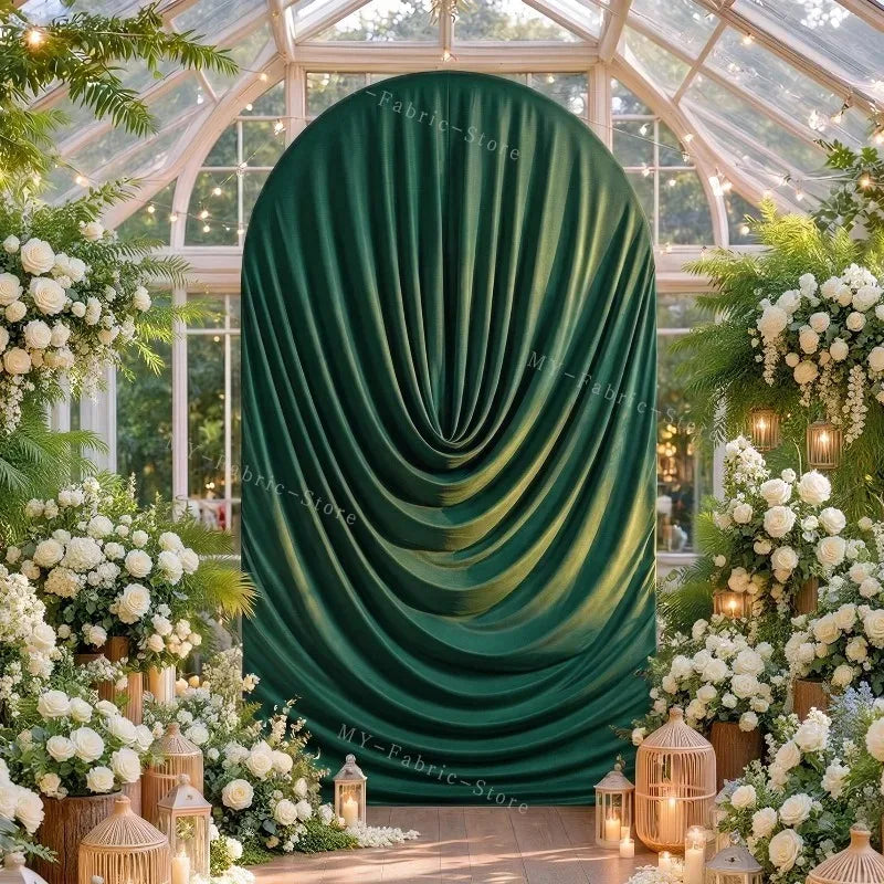 A deep emerald green fabric draped in pleats serves as an arch cover for a wedding or event setting surrounded by white flowe