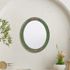 An oval wall mirror with a green frame featuring intricate lace design mounted on a white wall.