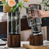 A clear glass vase with a wooden base holding green plants, being held by a hand.