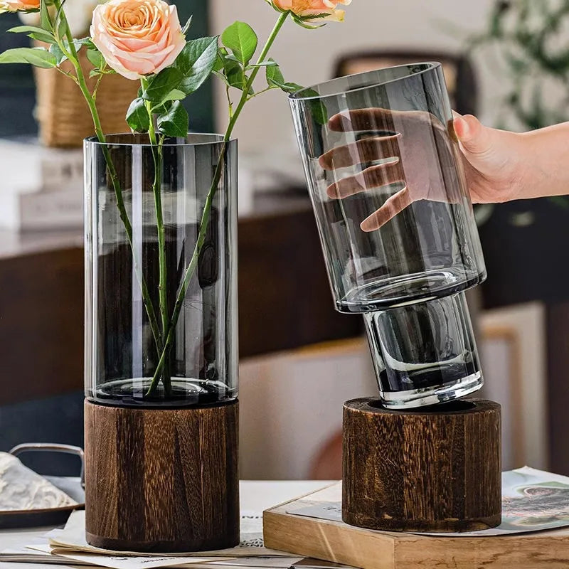 A clear glass vase with a wooden base holding green plants, being held by a hand.