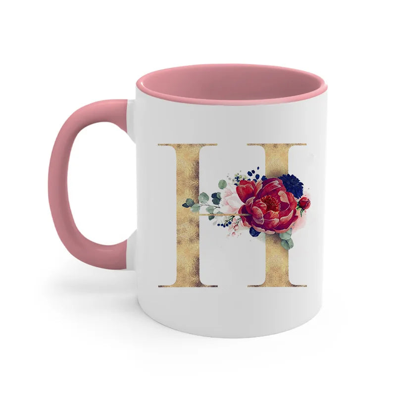 A pink ceramic coffee mug featuring a gold letter initial decorated with a floral arrangement.