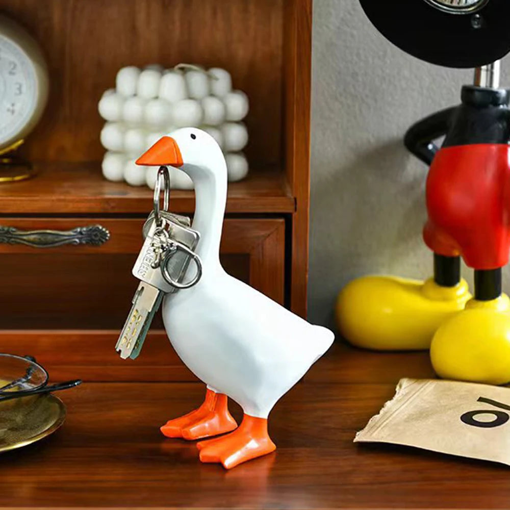 A white ceramic goose key holder with an orange beak and feet sits on a wooden surface.