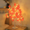 An LED maple leaf table lamp with warm glow illuminates orange-red leaves on a wooden surface.