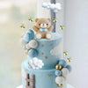 A two-tiered light blue cake decorated with clouds, stars, and a teddy bear topper.