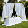 A white fabric backdrop curtain decorated with floral arrangements set up outdoors for an event.