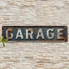 A weathered, dark metal sign with the word GARAGE mounted on a light-colored stone wall.