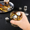 A hand holds a glass of amber liquid with reusable stainless steel ice cubes on a dark surface.