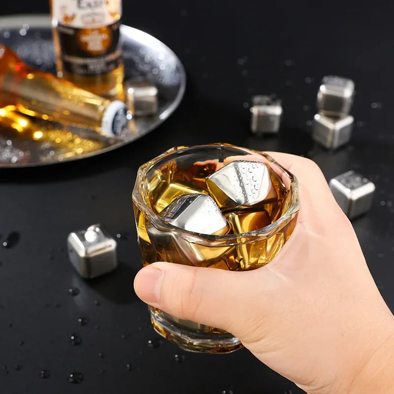 A hand holds a glass of amber liquid with reusable stainless steel ice cubes on a dark surface.