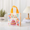 A pink and yellow non-woven gift tote bag with a birthday print and orange handles sits on a white surface.