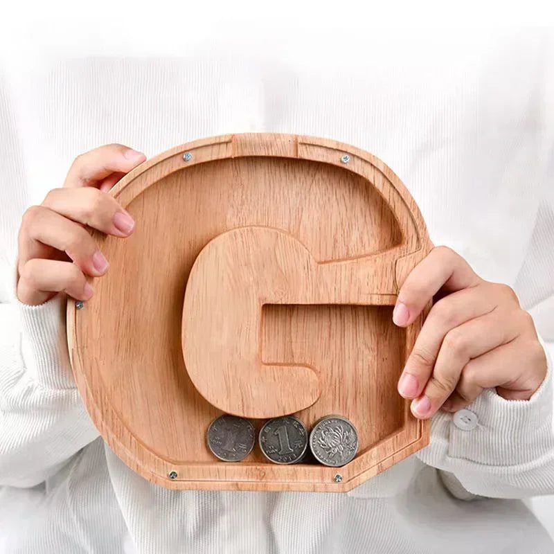 A child's hands hold a wooden alphabet piggy bank coin box with a letter G carved into it.