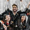 Four smiling graduates in black gowns and caps pose against a silver metallic foil fringe curtain backdrop.