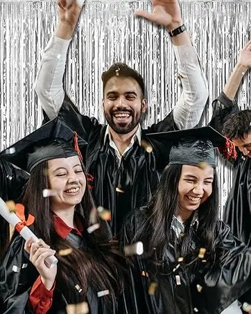 Four smiling graduates in black gowns and caps pose against a silver metallic foil fringe curtain backdrop.