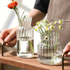A hand holds a clear glass vase with vertical stripes filled with fresh flowers on a wooden surface.