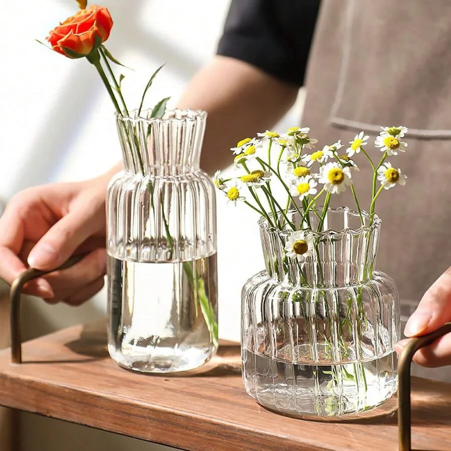 A hand holds a clear glass vase with vertical stripes filled with fresh flowers on a wooden surface.