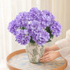 A bouquet of purple artificial hydrangea silk flowers in a patterned vase on a wooden table.