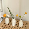 Three white ceramic mini flower vases holding green leafy stems on a wooden tray.