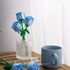 A bouquet of blue roses made from DIY building blocks displayed in a glass vase next to a gray mug.