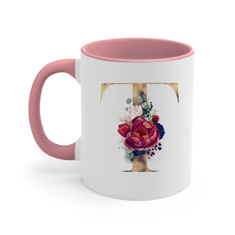 A pink ceramic coffee mug featuring a gold letter T design with floral accents.