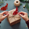 Hands tie a brown gift box with red artificial holly berry stems for Christmas decor