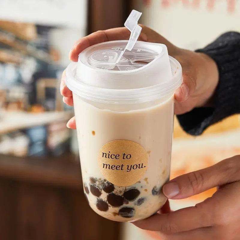 A hand holds a clear disposable coffee cup filled with a light-colored beverage and dark pearls, featuring a lid.