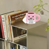 A pink crocheted tea cup ornament with a motivational charm sits on a shelf among books and greenery.