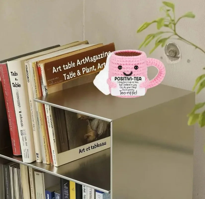 A pink crocheted tea cup ornament with a motivational charm sits on a shelf among books and greenery.