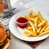 A plate of french fries and a small cup of red sauce on a white plate with newspaper in the background.